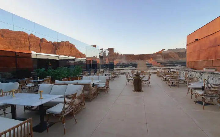 A modern outdoor dining terrace with white seating featuring a massive mirrored wall reflecting the red desert canyons of AlUla.