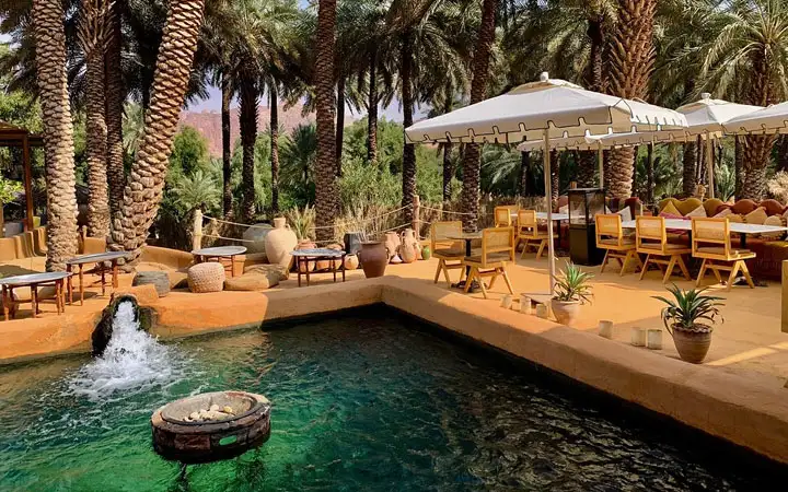 A tranquil outdoor restaurant nestled among tall palm trees featuring an emerald green pool with a fountain and traditional clay pottery decor.