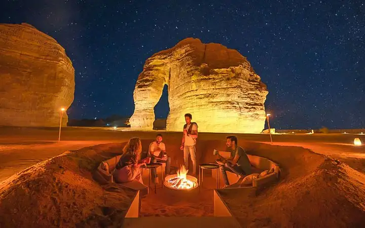 A group of people sitting in a sunken fire pit at night, illuminated by a warm fire with the giant Elephant Rock formation and a starry sky in the background.